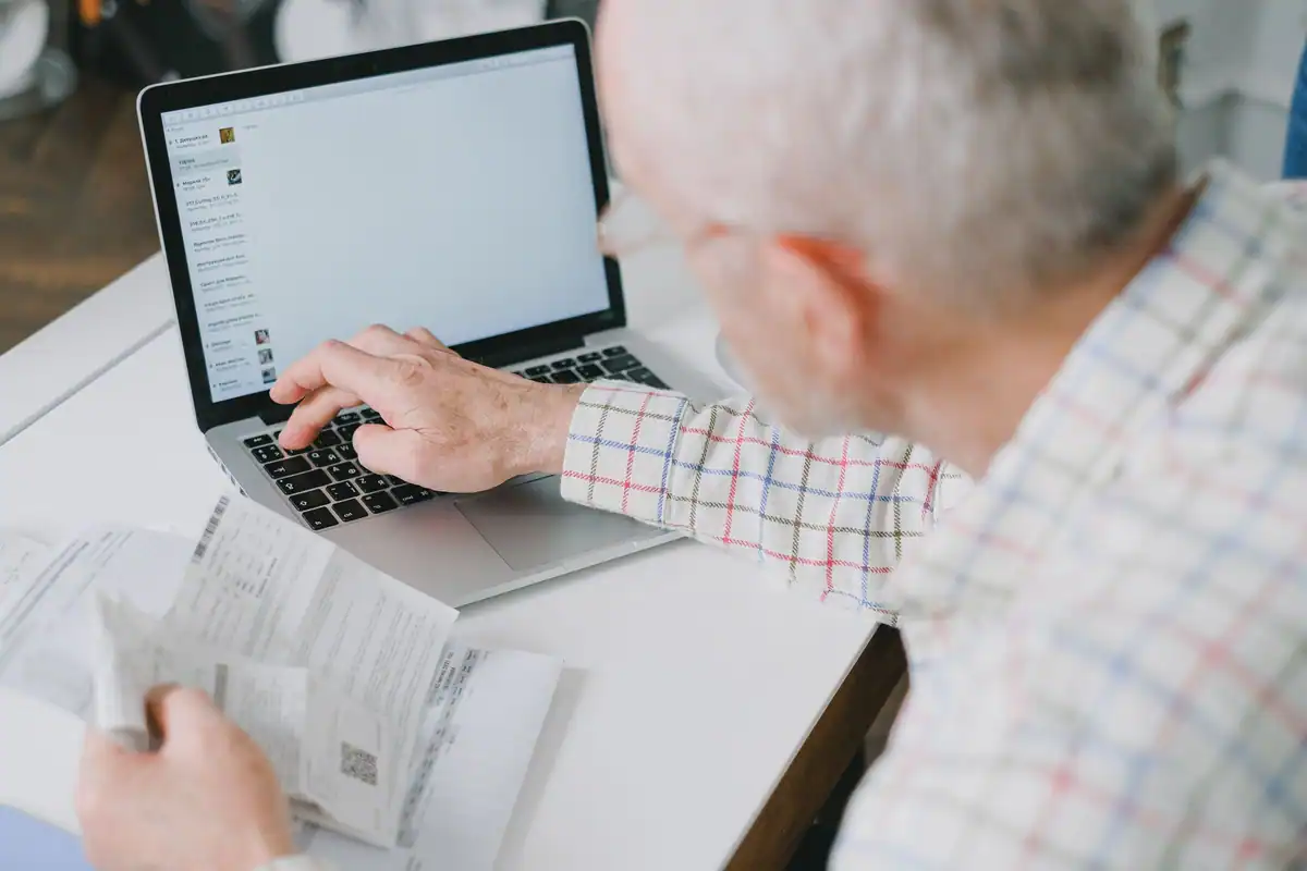 Bărbat în vârstă la laptop, cu o foaie în mână, verificând informațiile online