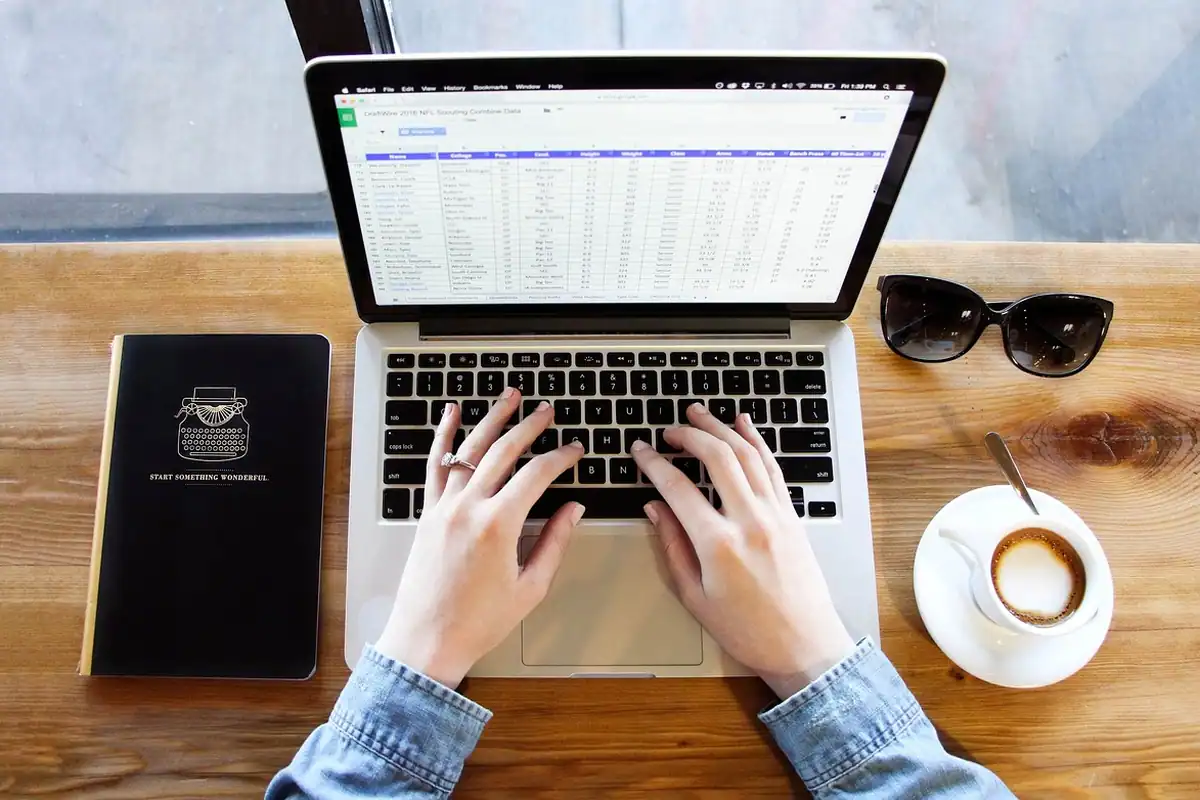 Mâini care tastează pe un laptop, lângă o ceașcă de cafea și ochelari de soare