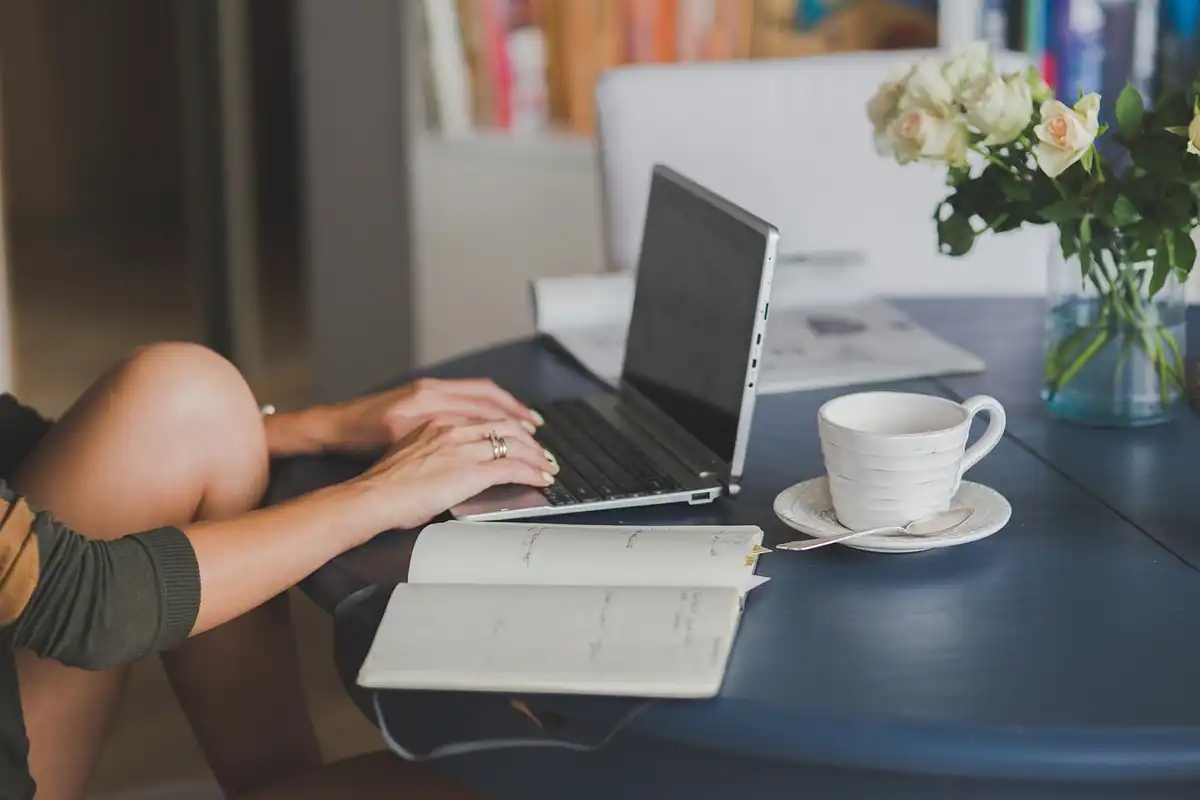 Femeie lucrează la laptop, cu un caiet și o ceașcă de cafea alături