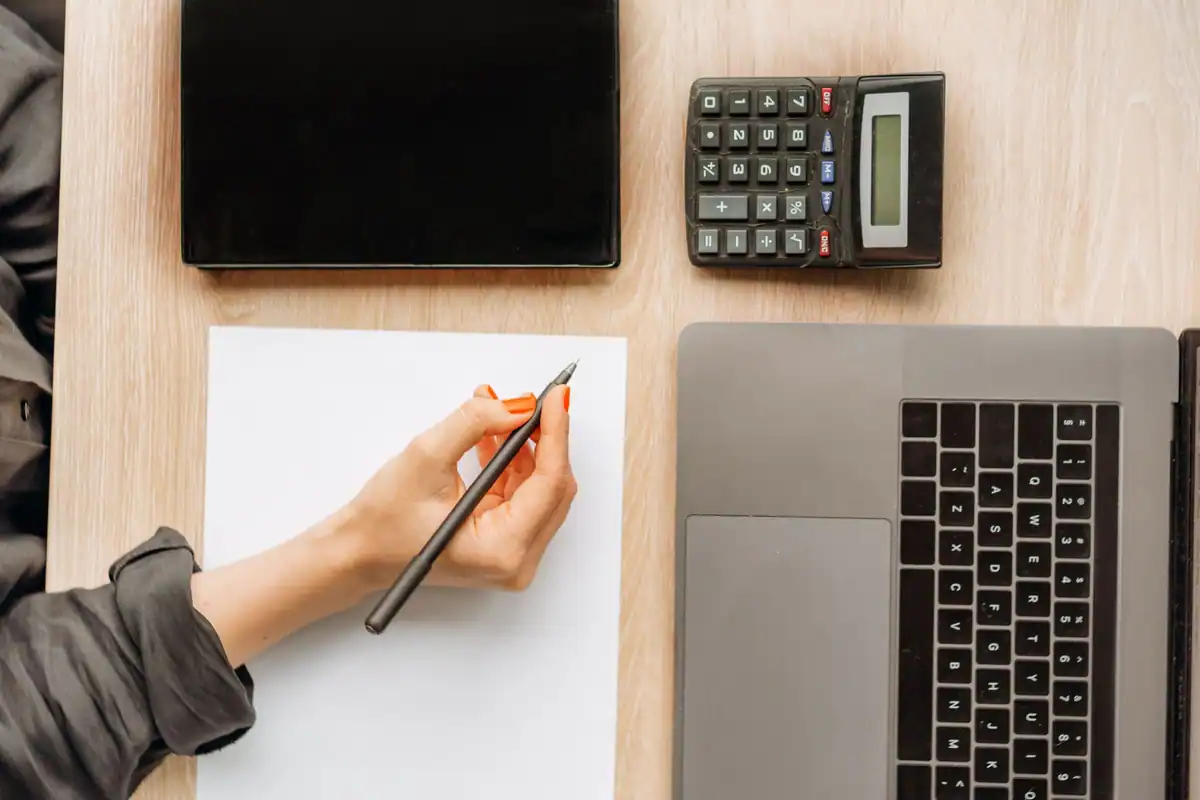 Mână care notează pe o foaie, lângă laptop, tabletă și calculator