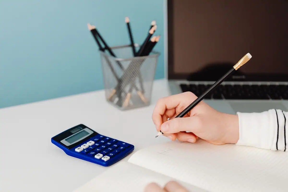 Mână care scrie într-un caiet, lângă un calculator albastru și un pahar cu pixuri
