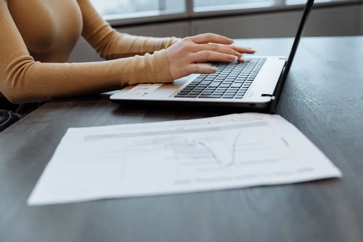 Mâini care tastează pe un laptop, lângă o foaie cu un grafic